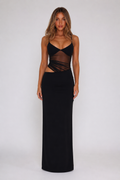 A woman modeling a black sleeveless maxi dress with a sheer mesh crossover bodice and side cutouts, photographed against a plain light gray studio background.